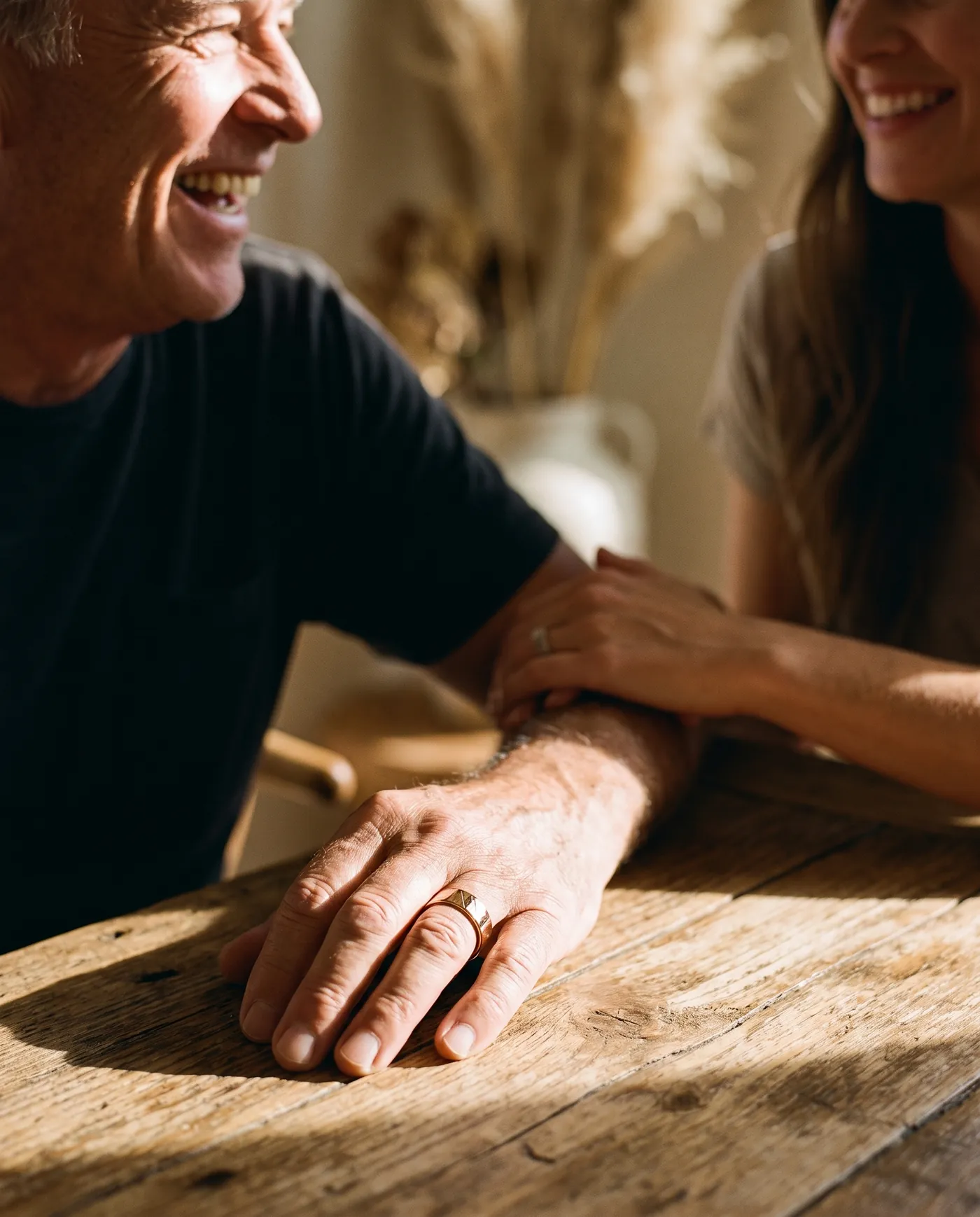 A father and his adult daughter laughing at a sunlit table, his hand wearing the ring